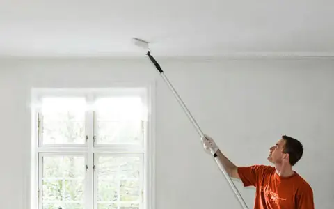 Man målar taket med en lång roller. Fönstret bakom släpper in ljus.