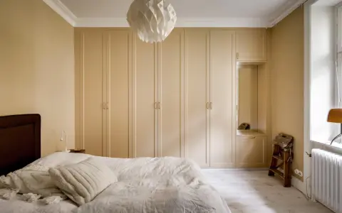 Sovrum med en stor, ljusbeige garderob som sträcker sig längs väggen. En vit taklampa med geometriskt mönster hänger ovanför.