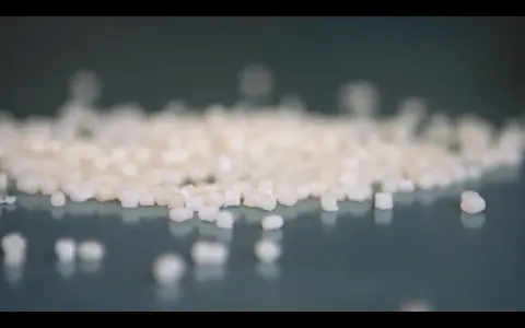 Små vita granulatkorn, råmaterial för nedbrytbara plastpåsar, ligger utspridda på en mörk yta.