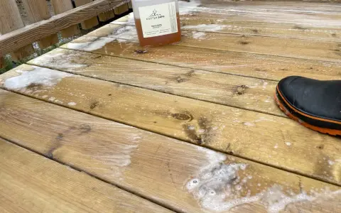 En flaska linoljesåpa står på en altan som håller på att rengöras. En svart stövel med orange kant syns i bildens nedre högra hörn.