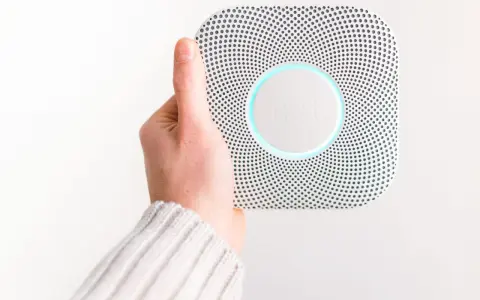 En hand håller upp en vit Google Nest Protect-rökdetektor med blåaktig glöd.