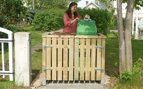 En kvinna lägger sopor i en grön tunna inbyggd i ett trähus. Ett smart sätt att bygga in soptunnor i trädgården.
