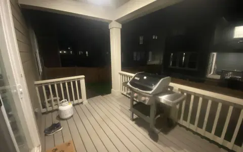 En gasolgrill under taket på altanen, redo för grillkvällar. Bredvid står en gasoltub och ett par skor.