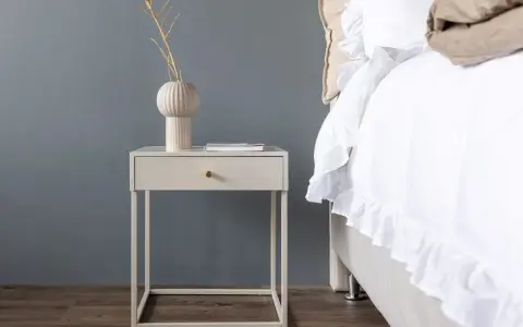 Ett stilrent sängbord i beige med en låda och en vas med kvistar. Bredvid står en inbjudande säng med vitt, volangprytt överkast.
