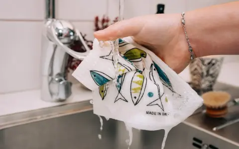 En hand håller en disktrasa i linne med fiskmönster under rinnande vatten i ett kök.