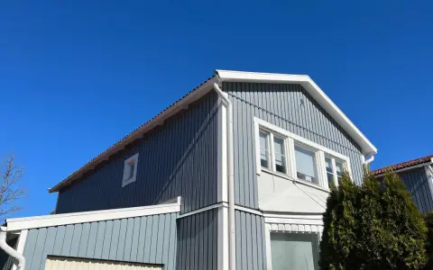 Blå himmel över ett gråblått trähus. Snart dags att måla fasad för att skydda det mot väder och vind.