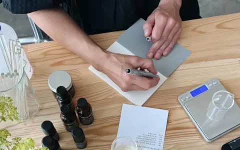 Person skriver recept för ekologiska doftpinnar. Ingredienser och verktyg ligger på ett träbord.