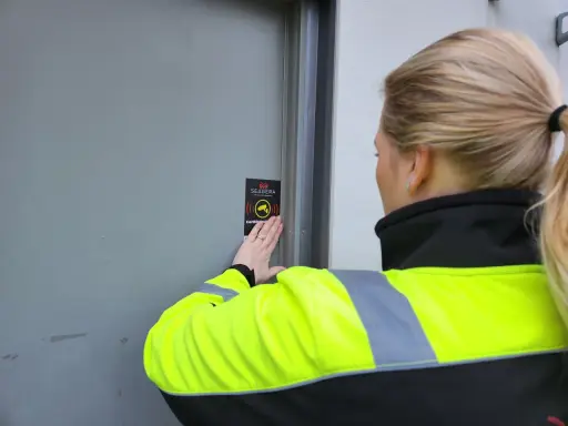En person med reflexväst fäster en skylt med texten "Kameraövervakning" på en dörr, vilket indikerar ett larm på en byggarbetsplats.