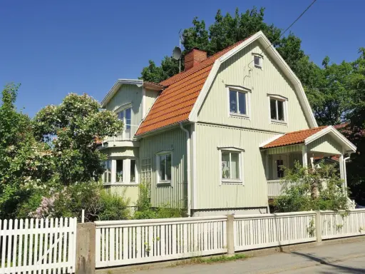 Ljusgrönt trähus med tegeltak och vit staket. En vacker trädgård med blommande buskar. Planerar att byta fasad för att fräscha upp huset.