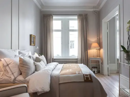 Ett elegant sovrum med en stor säng bäddad med vita lakan och en brun pläd med H-mönster. Vackra möbler till sovrummet som ett litet sidobord med lampa och en orkidé.