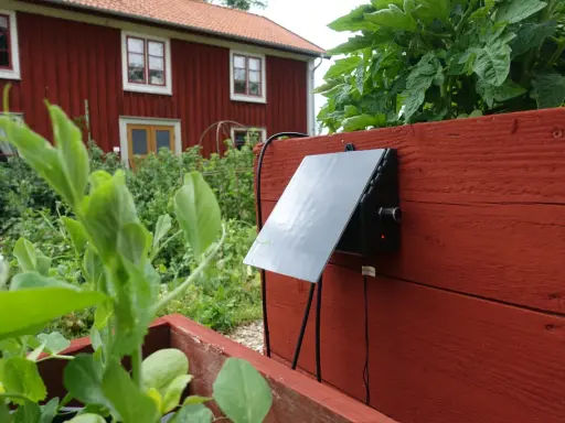 En solcellsdriven bevattning är monterad på en röd träkista i en trädgård, med ett rött hus i bakgrunden.
