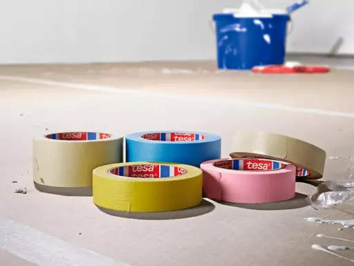 Fem rullar maskeringstejp i olika färger ligger på ett golv som täckts med papper. En blå hink och en röd roller syns i bakgrunden.