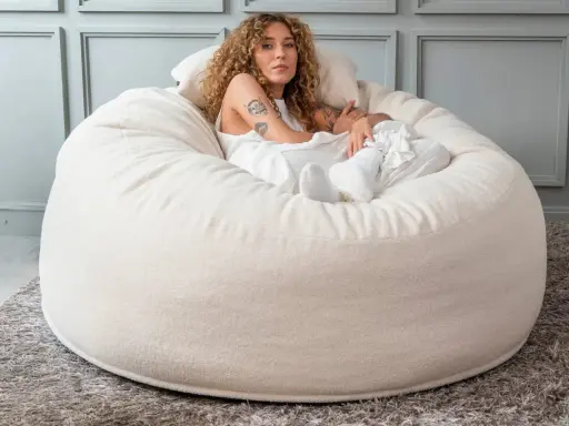 En kvinna med lockigt hår vilar i en stor, mjuk beige sittpall.