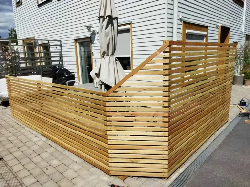 Nybyggt staket i trä ramar in uteplatsen. Grillen och parasollet syns bakom.