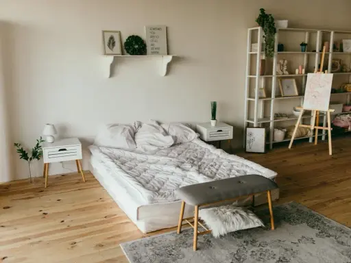 Ett sovrum med en stor, vit madrass som ser ut att vara förvaring under. En hylla med dekorationer, ett staffli med en tavla och en bokhylla fyller rummet.