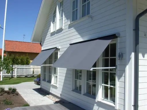 Grå markiser monterade på ett vitt trähus. Solen skiner och skapar skuggor på fasaden.