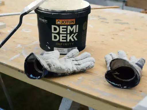 Förberedelser för målning utomhus: en burk Jotun Demi Dekk, pensel och slitna handskar ligger på ett träbord.