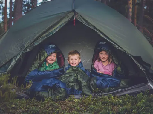Tre glada barn myser i sina sovsäckar i ett tält. En perfekt tältsäng barn för äventyr i skogen.