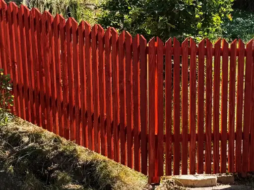 Rött, spetsigt staket av tryckimpregnerat trä. Solen lyser genom plankorna och skapar skuggor.
