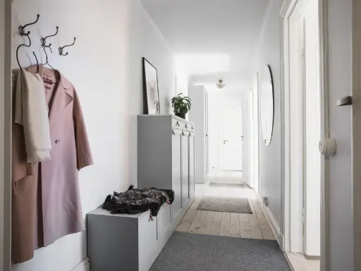 En smal hall med en rosa kappa och en vit halsduk på krokar. En grå förvaringsmöbel med en fårskinnspläd.