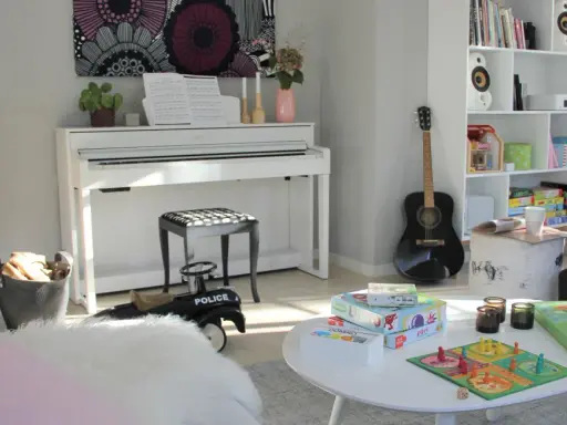 Ett mysigt matrum med en ståtlig kamin, ett vitt piano och en bokhylla fylld med böcker och leksaker.