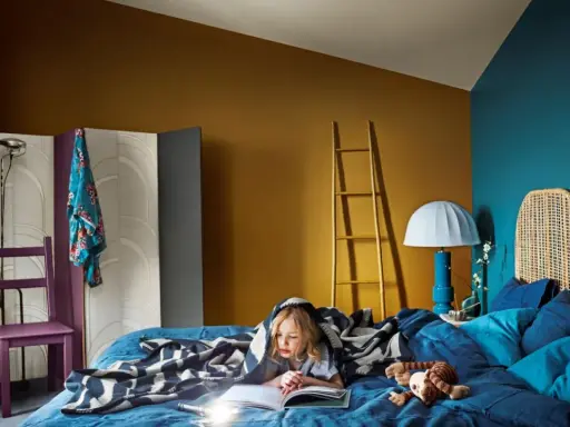 Ett barn läser under ett mönstrat täcke i ett mysigt blått sovrum. En golvlampa och en stege i trä bidrar till rummets unika atmosfär.