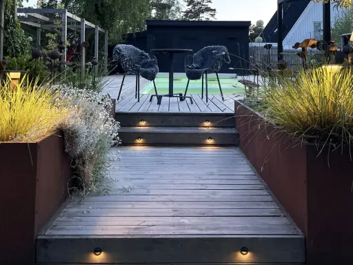 Välkomnande altan med decklights som lyser upp trappstegen. Två mysiga fåtöljer väntar vid ett runt bord.