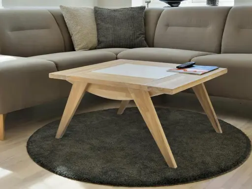 Ett smart soffbord i ljust trä med en vit inläggning, placerat på en rund, mörk matta framför en beige hörnsoffa.