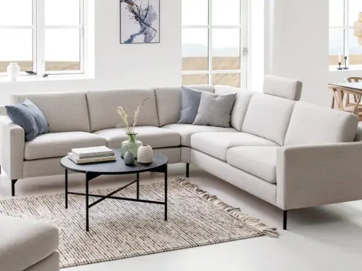 Ljusbeige hörnsoffor i ett modernt vardagsrum med ett runt soffbord, en matta och stora fönster.