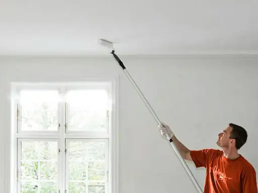 Man målar taket med en lång roller. Fönstret bakom släpper in ljus.