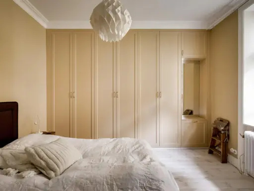 Sovrum med en stor, ljusbeige garderob som sträcker sig längs väggen. En vit taklampa med geometriskt mönster hänger ovanför.