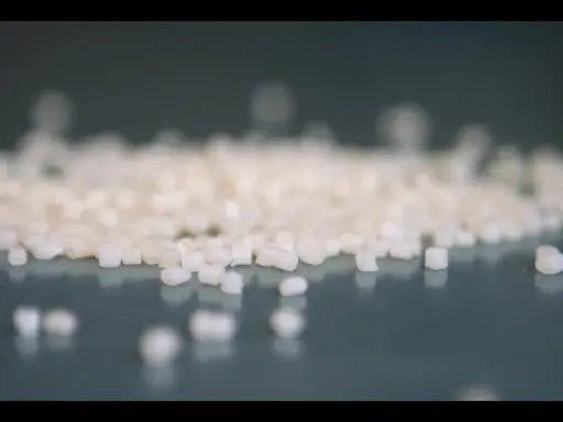 Små vita granulatkorn, råmaterial för nedbrytbara plastpåsar, ligger utspridda på en mörk yta.