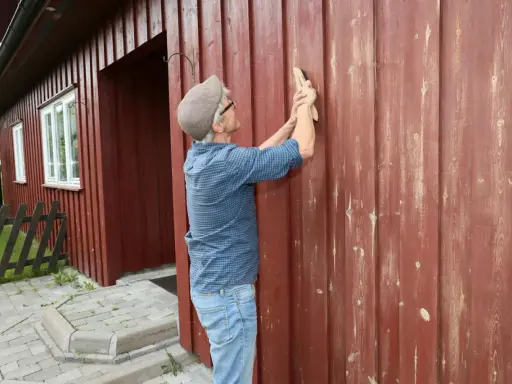 Man slipar träfasaden på ett rött hus. Förberedelser för att måla om.