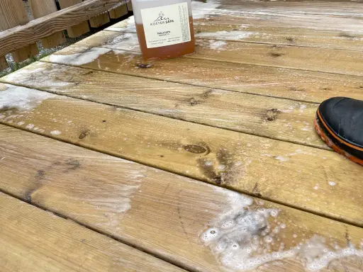 En flaska linoljesåpa står på en altan som håller på att rengöras. En svart stövel med orange kant syns i bildens nedre högra hörn.