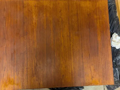 En träbordsskiva ligger på svart plast. Bredvid ligger en slipmaskin och en trasa, redo att måla bordsskivan.