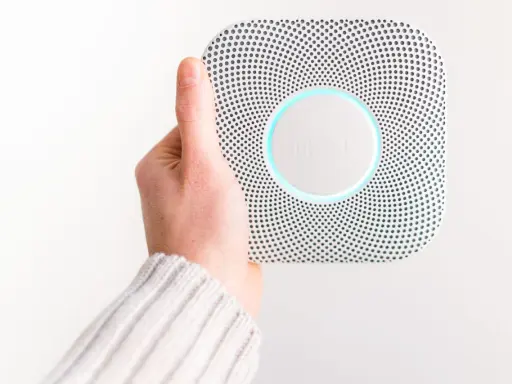 En hand håller upp en vit Google Nest Protect-rökdetektor med blåaktig glöd.
