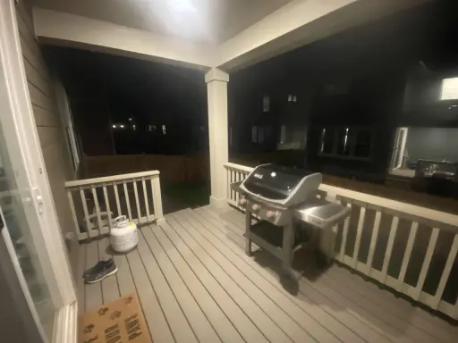 En gasolgrill under taket på altanen, redo för grillkvällar. Bredvid står en gasoltub och ett par skor.