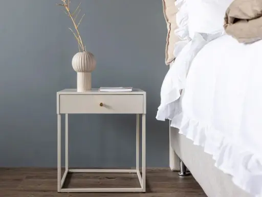 Ett stilrent sängbord i beige med en låda och en vas med kvistar. Bredvid står en inbjudande säng med vitt, volangprytt överkast.