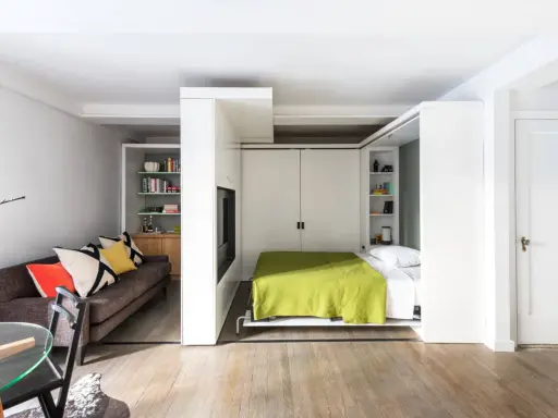 En smart säng i vardagsrummet som fälls ner från väggen. En mysig soffa med färgglada kuddar står bredvid.