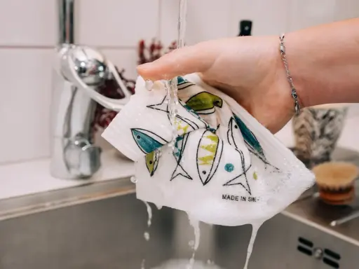 En hand håller en disktrasa i linne med fiskmönster under rinnande vatten i ett kök.