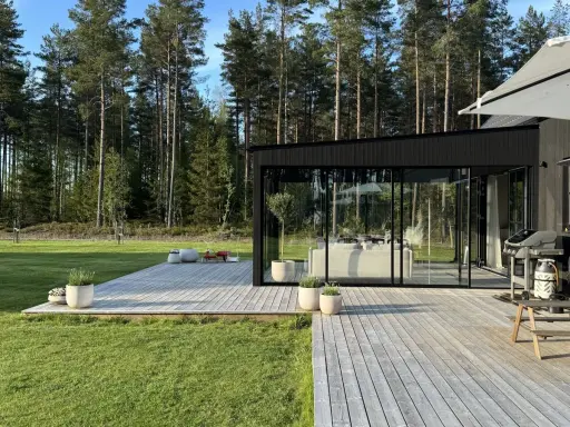 Enkel uteplats med trätrall och glaspartier som vetter mot en grönskande skog. Perfekt för avkoppling.