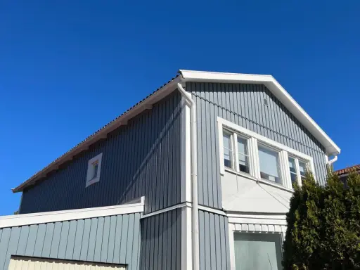 Blå himmel över ett gråblått trähus. Snart dags att måla fasad för att skydda det mot väder och vind.
