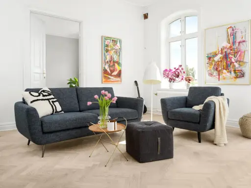 En modern soffgrupp 3+2 i mörkblått tyg, med en fåtölj, soffbord, pall och konst på väggarna.
