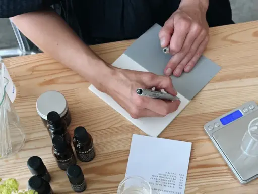 Person skriver recept för ekologiska doftpinnar. Ingredienser och verktyg ligger på ett träbord.