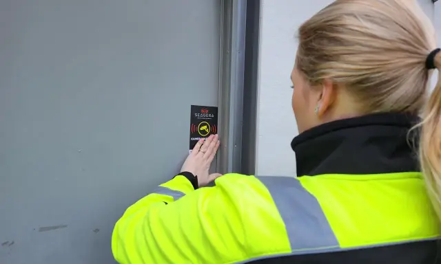 En person med reflexväst fäster en skylt med texten "Kameraövervakning" på en dörr, vilket indikerar ett larm på en byggarbetsplats.