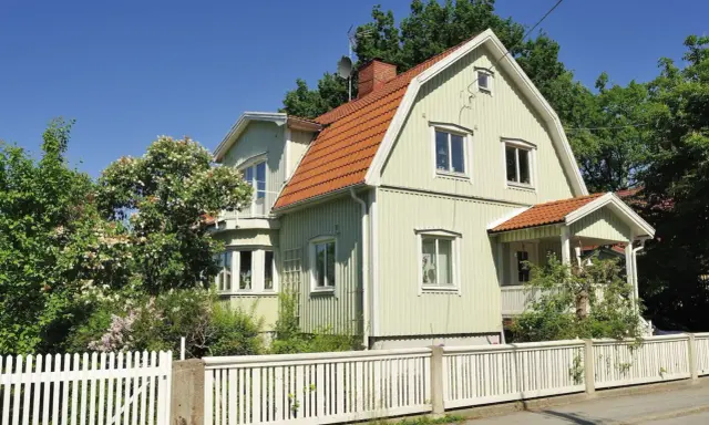Ljusgrönt trähus med tegeltak och vit staket. En vacker trädgård med blommande buskar. Planerar att byta fasad för att fräscha upp huset.