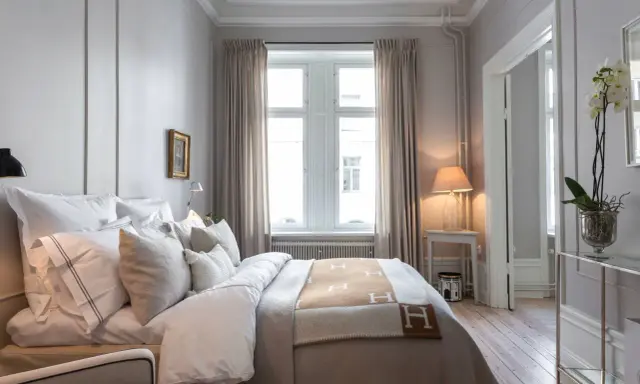 Ett elegant sovrum med en stor säng bäddad med vita lakan och en brun pläd med H-mönster. Vackra möbler till sovrummet som ett litet sidobord med lampa och en orkidé.