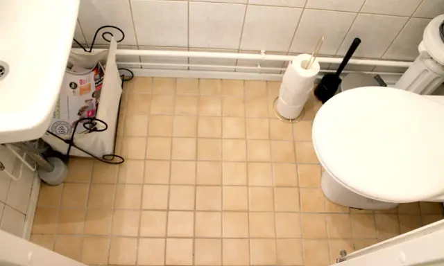 En liten toalett med beige kakelgolv. En tidningskorg står vid handfatet, och en toalettrulle och borste står bredvid toaletten. Kanske dags att måla kakel?