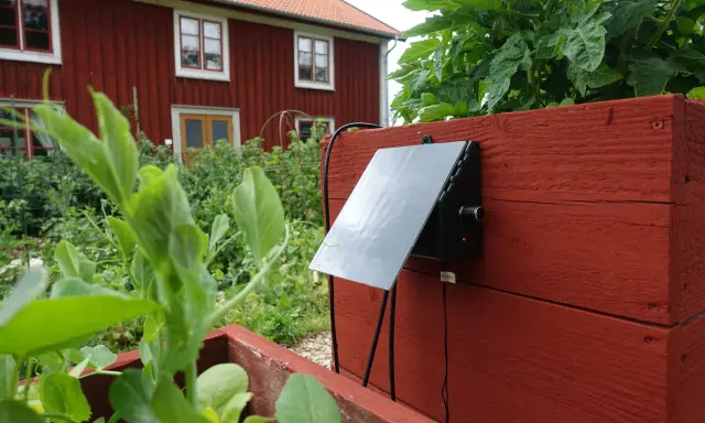 En solcellsdriven bevattning är monterad på en röd träkista i en trädgård, med ett rött hus i bakgrunden.
