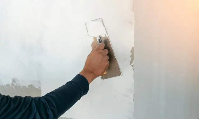 Handen sprider spackel på en vägg med en bredspackel. Arbetet med att måla på spackel fortsätter för att skapa en jämn yta.